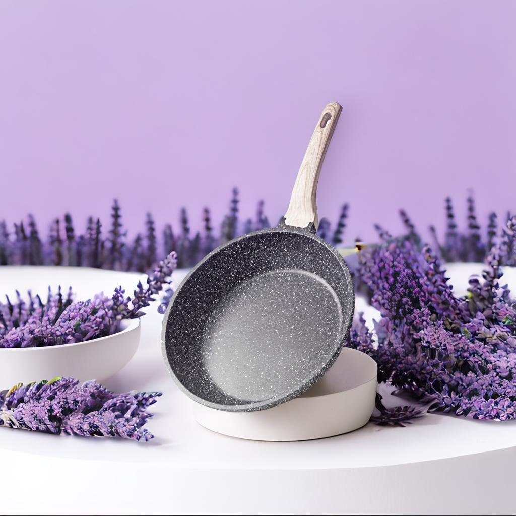 A non-stick gray frying pan with a wooden handle is displayed on a white surface surrounded by fresh lavender flowers against a soft purple background.